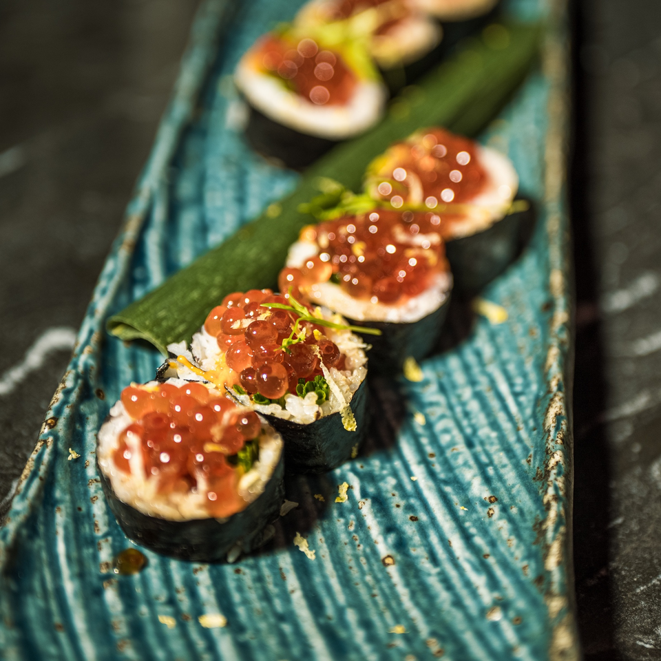 Close-up of sushi rolls topped with orange fish roe, cream filling, herbs, and green leaves on a rustic blue platter.