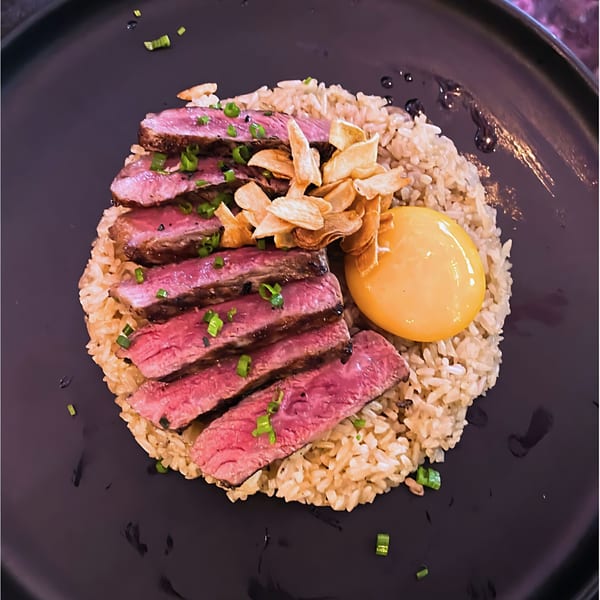 Sliced steak with rice, crispy garlic, and a raw egg yolk on a dark plate.
