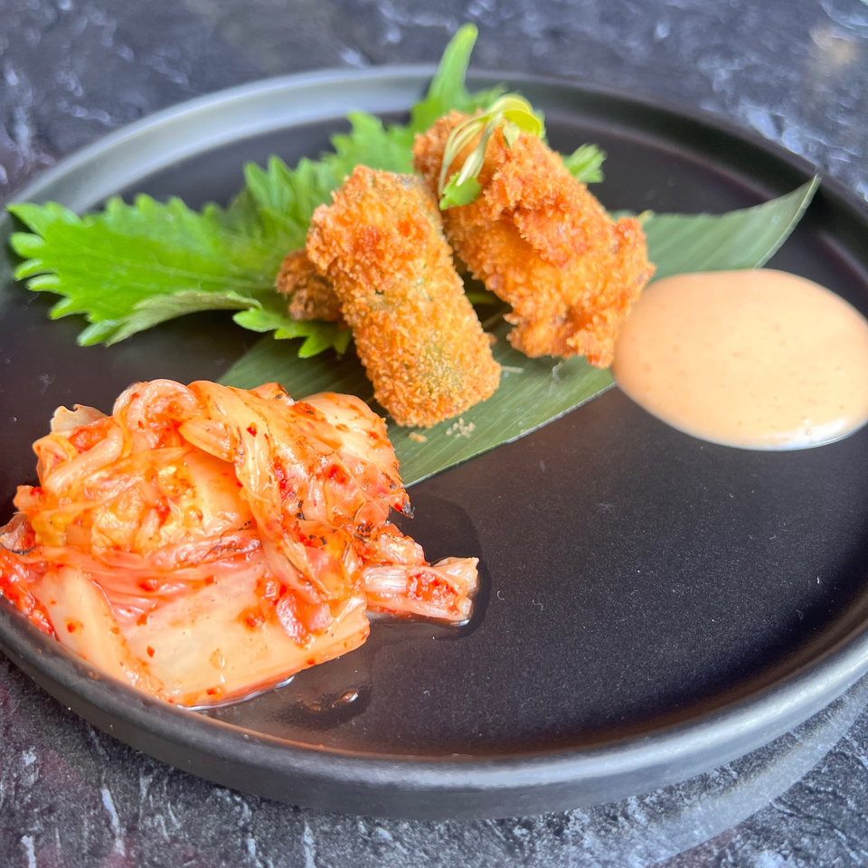 Crispy fried appetizers with kimchi, greens, and creamy dipping sauce arranged on a black plate.