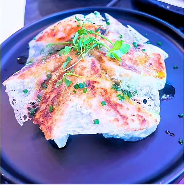 Crispy gyoza with delicate lace-like edges, garnished with fresh pea shoots and chives on a dark plate.