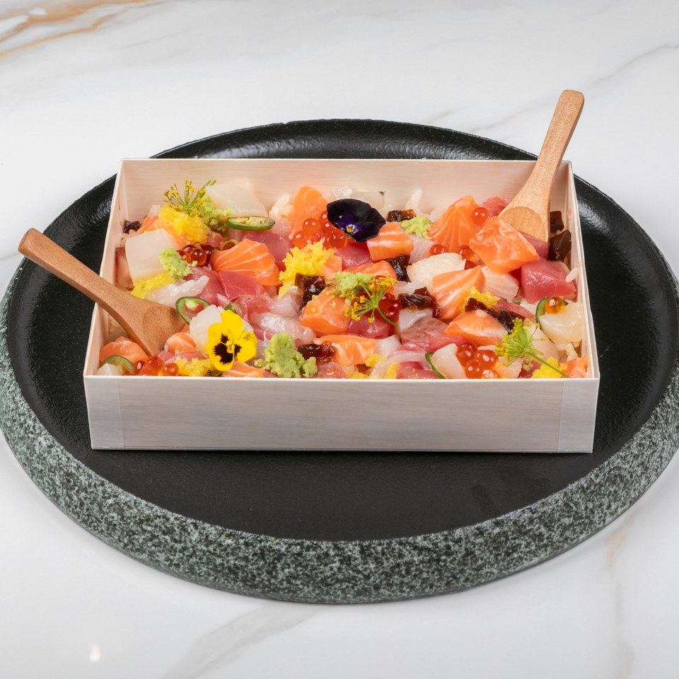 Colorful sashimi assortment with salmon, tuna, vegetables, edible flowers, and garnishes in a wooden box on a black platter.