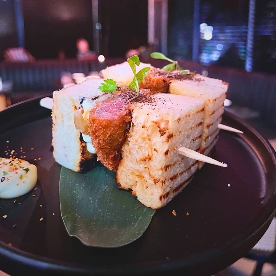 Toasted sandwich wedges with creamy filling, a golden croquette, herbs, and dipping sauce on a dark plate.