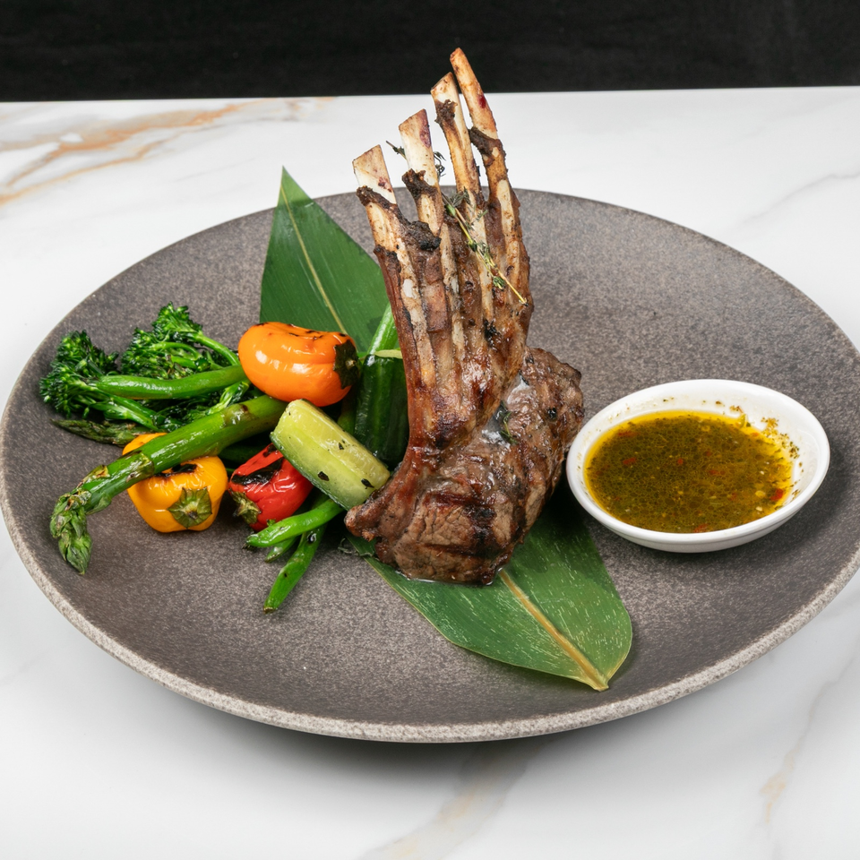 Grilled rack of lamb with roasted vegetables, tomatoes, leafy garnish, and dipping sauce on a dark ceramic plate.